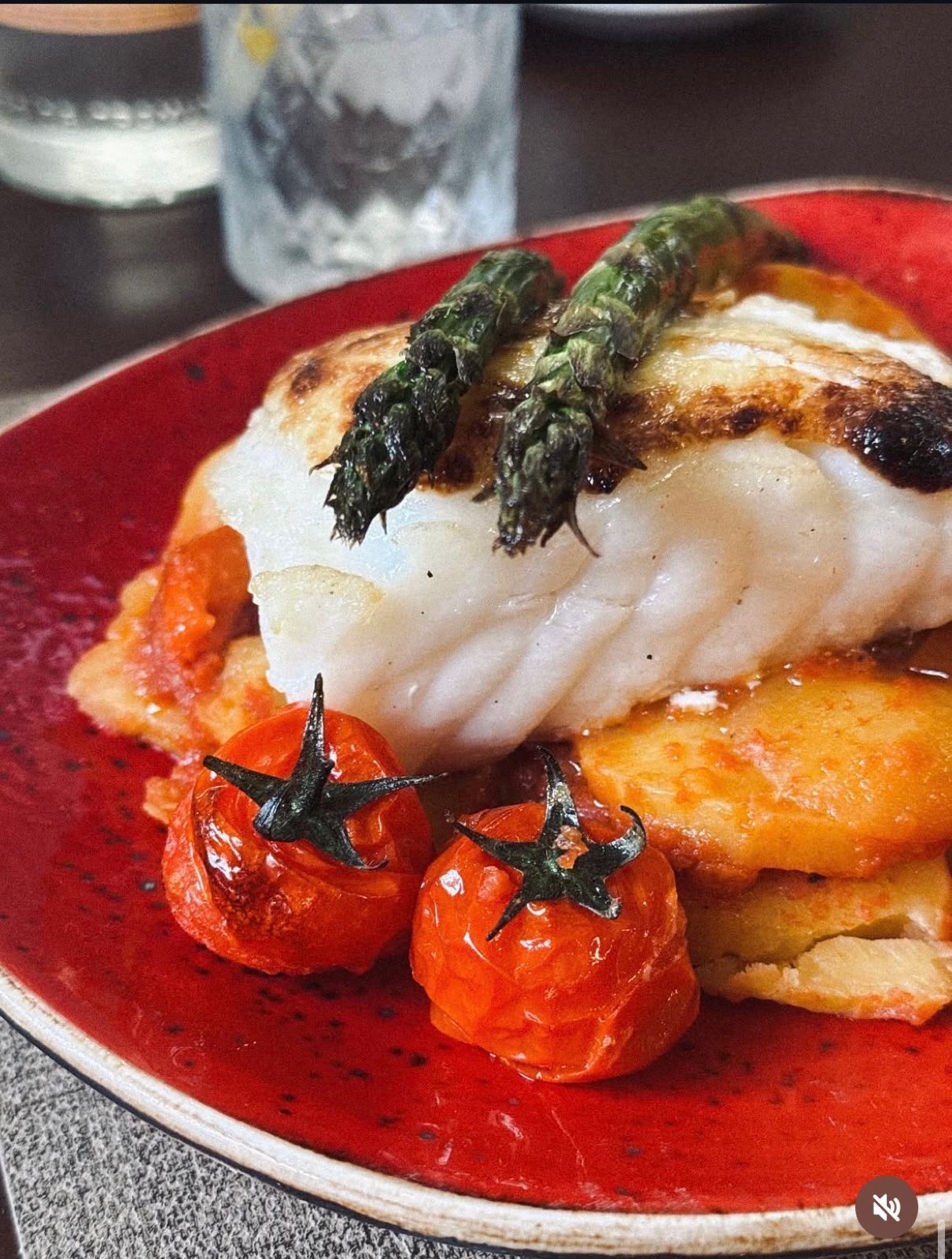 Bacalao Jumbo al horno con espárragos trigueros, tomates cherry, una cama de tumbet (patata, berenjena y calabacín) y alioli gratinado, en un plato rojo recién salido del horno.